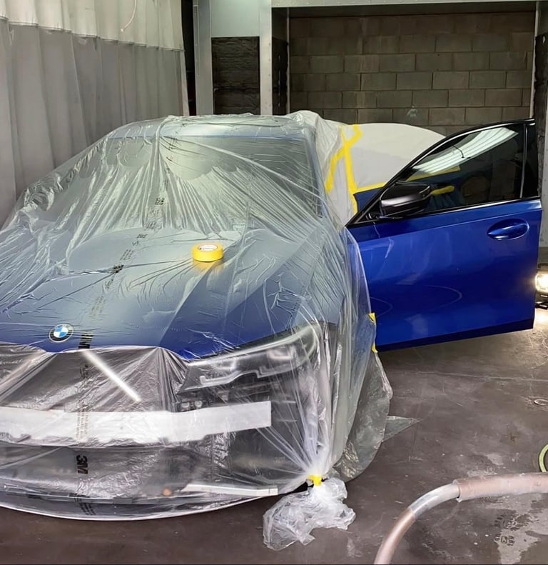 Blue car covered in clear plastic sheeting during paint or protection work in an indoor garage